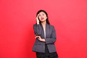 The expression of an Asian office woman with her arms crossed, dizzy, has a headache, while holding the camera, wearing a gray jacket and skirt with a red background. for health and advertising photo