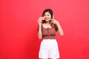 gesture of a smiling Asian woman holding a debit credit card from the side while pointing at it, wearing a brown blouse with a red background. for transaction, technology and advertising concepts photo