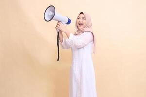 young girl asian wearing hijab slanted to the right candidly shouts cheerfully holding a loudspeaker megaphone with both hands. Technology, broadcast and promotion concept photo