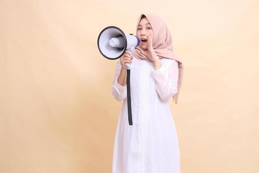 girl asian young wearing a hijab at the camera shouts gracefully holding a loudspeaker megaphone while focusing the sound. Technology, broadcast and promotion concept photo