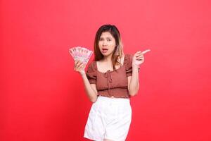 expression of a sad indonesia woman holding rupiah money while pointing to the left wearing a brown blouse top with a red background. for transaction, technology and advertising concepts photo