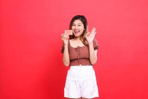 gesture of a cheerful indonesia woman holding a debit credit card with a zero sign okay, good wearing a brown blouse with a red background. for transaction, technology and advertising concepts photo