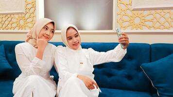 portrait of a pair of beautiful Asian women sitting on the sofa smiling gracefully selfie in front of the cellphone camera with an elegant Arabic nuance in the background of Islamic calligraphy photo