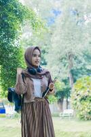 mature asian woman in veil candid smile carrying books and clipboard and bag standing in outdoor park. female students for the themes of education, technology and advertising photo