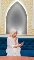 beautiful Asian woman wearing a hijab with a very shocked expression checking her cell phone gadget sitting on the sofa with a background of Islamic ornamental calligraphy in Arabic nuances photo