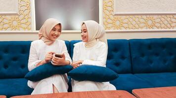 Two young Asian women with cheerful smiles are discussing with a background of Islamic ornamental calligraphy with elegant Arabic nuances sitting on the sofa photo
