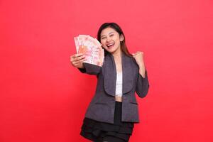 the expression of a cheerful indonesia director woman holding a credit card and money in front of her clenched fist at the camera wearing a jacket and skirt on a red background. for business advert photo