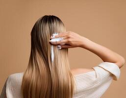 Young woman applying hair mask on beige background, back view photo