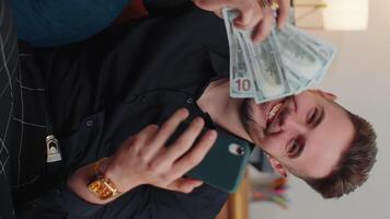 Smiling happy young bearded man counting money cash use smartphone, income, saves, lottery win home video