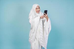 Attractive Asian Muslim woman in prayer gown using phone to text or browse internet, isolated by blue background. Hajj concept. photo
