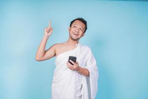 Joyful Asian Muslim man in ihram holding a smart phone while pointing to the copy space upwards, isolated by blue background. Hajj concept. photo