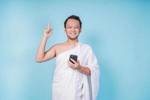 Joyful Asian Muslim man in ihram holding a smart phone while pointing to the copy space upwards, isolated by blue background. Hajj concept. photo