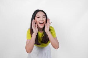Young Asian woman in a yellow shirt is making a whispering gesture with hands on her face, isolated by white background. Concept of surprise or shock. photo