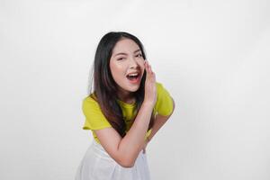 Young Asian woman in a yellow shirt is making a whispering gesture with hands on her face, isolated by white background. Concept of surprise or shock. photo