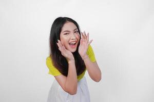 Young Asian woman in a yellow shirt is making a whispering gesture with hands on her face, isolated by white background. Concept of surprise or shock. photo