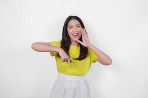 Excited young Asian woman wearing yellow shirt and white skirt making shout gesture while pointing to the copy space downwards, isolated by white background. photo