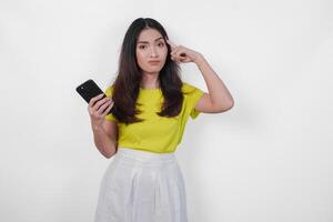 Thoughtful Asian woman in a yellow shirt is holding a cell phone in her hand over isolated white background. photo