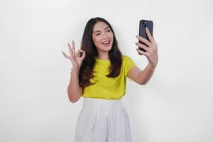 Happy young Asian woman in a yellow shirt is taking a selfie with her cell phone. She is smiling and showing okay good finger gesture over isolated white background. photo