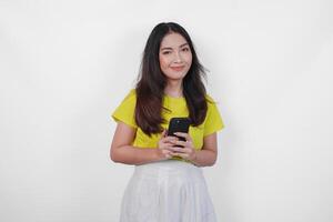 Cheerful Asian woman in a yellow shirt is holding a cell phone, smiling to the camera feeling happy, isolated by white background. photo