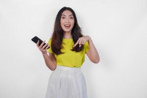 Smiling young woman is holding a cell phone and pointing to the copy space downwards over isolated white background. photo