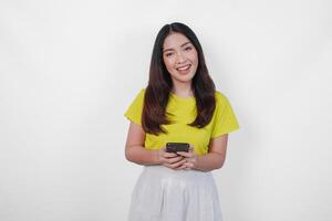 Cheerful Asian woman in a yellow shirt is holding a cell phone, smiling to the camera feeling happy, isolated by white background. photo