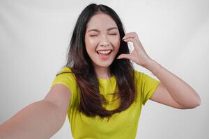 Cute young Asian woman in a yellow shirt is taking a selfie while making a heart gesture on her cheek, isolated over white background. photo
