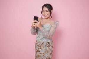 Cheerful Asian woman wearing modern kebaya using phone to text or browse internet, isolated by pink background. photo