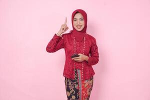 Joyful Asian woman wearing modern kebaya dress and hijab pointing at the copy space upward while holding smartphone, isolated by pink background. photo