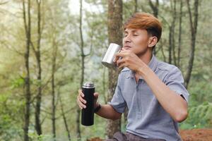 Portrait of excited Asian man pouring water from the vacuum flask to the stainless steel mug. Drinking mineral water while trekking in nature forest photo