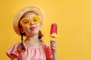 Cheerful little girl with fruit ice cream on yellow isolated background with copy space. Blonde female child with smirk on face in straw hat, sunglasses and pink top. She is holding a popsicle photo