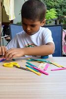 Smart Indian little boy performing best out of waste activity with different colourful objects during the summer vacations, Cute Indian Kid doing best our of waste activity at home on wooden table photo