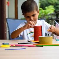 Smart Indian little boy performing best out of waste activity with different colourful objects during the summer vacations, Cute Indian Kid doing best our of waste activity at home on wooden table photo