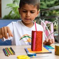 Smart Indian little boy performing best out of waste activity with different colourful objects during the summer vacations, Cute Indian Kid doing best our of waste activity at home on wooden table photo