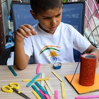 Smart Indian little boy performing best out of waste activity with different colourful objects during the summer vacations, Cute Indian Kid doing best our of waste activity at home on wooden table photo
