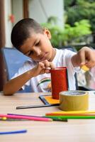 Smart Indian little boy performing best out of waste activity with different colourful objects during the summer vacations, Cute Indian Kid doing best our of waste activity at home on wooden table photo