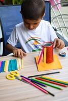Smart Indian little boy performing best out of waste activity with different colourful objects during the summer vacations, Cute Indian Kid doing best our of waste activity at home on wooden table photo
