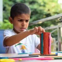 Smart Indian little boy performing best out of waste activity with different colourful objects during the summer vacations, Cute Indian Kid doing best our of waste activity at home on wooden table photo