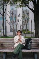 Woman sitting on a park bench smiling while using a smartphone with a backpack. Concept of outdoor relaxation and digital connectivity photo