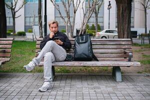 Man sitting on a park bench using smartphone with backpack and camera. Concept of urban relaxation and technology photo