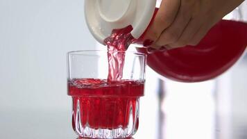 Female hand with red manicure. Red strawberry natural juice pouring in drinking glass glass from transparent pitcher in pink neon. Pouring red drink from a jug video
