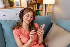 Woman listening music in headphones on windowsill background. Beautiful girl in denim and headphones is listening to music using a smart phone and smiling while lying on bed at home photo
