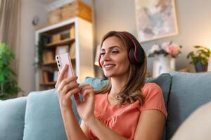 Woman listening music in headphones on windowsill background. Beautiful girl in denim and headphones is listening to music using a smart phone and smiling while lying on bed at home photo