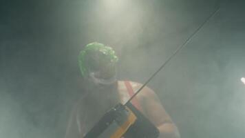 Scary man in the horror clown mask with green hair, standing in suspenders in the dark studio with flashing lights and threatening with the chainsaw close up video