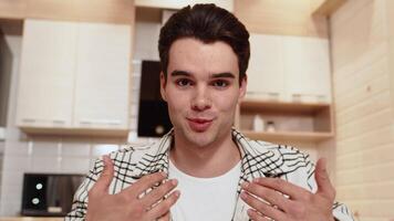 Front view of a young caucasian man in casual clothes sitting at home in a cozy kitchen and greetings his interlocutor via call at the beginning of a discussion video