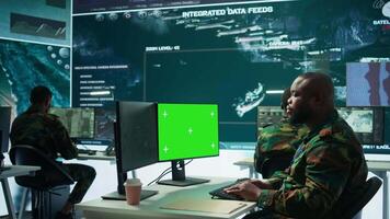Military specialist monitors satellite data next to green screen display, working at command post with high tech gear to support army defense operations. Coordinates strategic missions. Camera B. video
