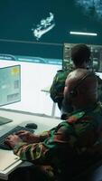 Vertical Military personnel in a base of operations using advanced computers and big screen, monitoring satellite systems to enhance security measures and process data. High tech army gear. Camera B. video