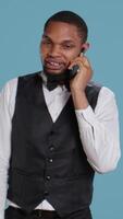 Vertical Hotel concierge in uniform makes room reservations on smartphone call, establishing lodging for clients at five star resort. Bellhop explaining amenities to guests in studio. Camera B. video