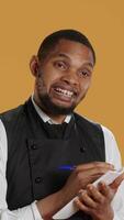 Vertical Professional caterer writing clients orders on notepad for the bill, takes notes of desired food against yellow background. Waiter with apron, notebook service for customer satisfaction. Camera A. video
