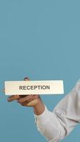 Vertical Hotel concierge staff showing the reception indicator at front desk, greeting guests at five star holiday resort in studio. Skilled professional bellboy holds sign against blue background. Camera A. video
