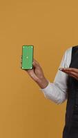 Vertical Restaurant staff showing a smartphone with green screen in studio, prepared to serve food orders in service oriented environment. Waiter with apron and uniform serving clients at tables. Camera B. video